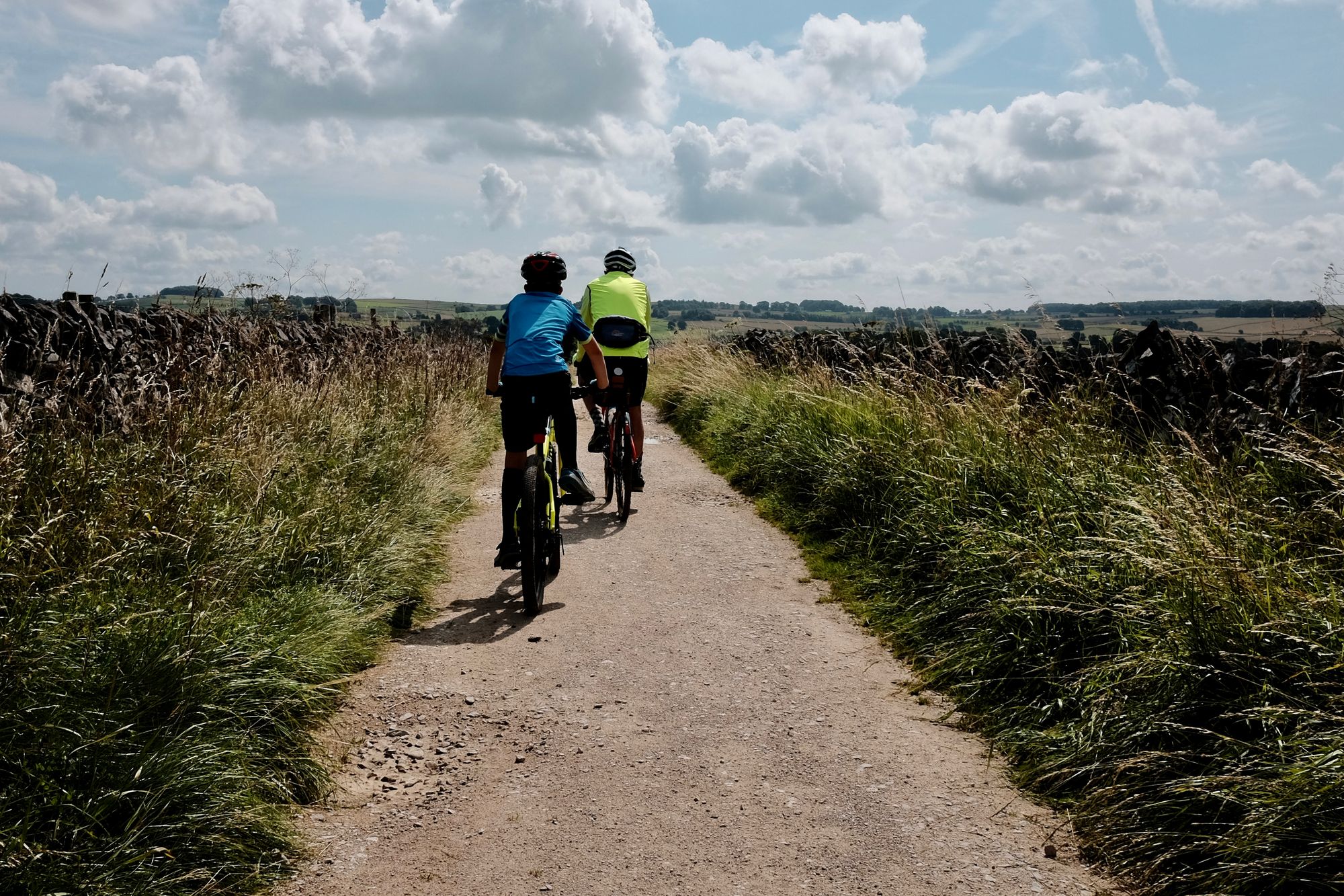 Two cyclists, a child in blue and an adult in fluro yellow, cycle away from the camera along a brown stone track.