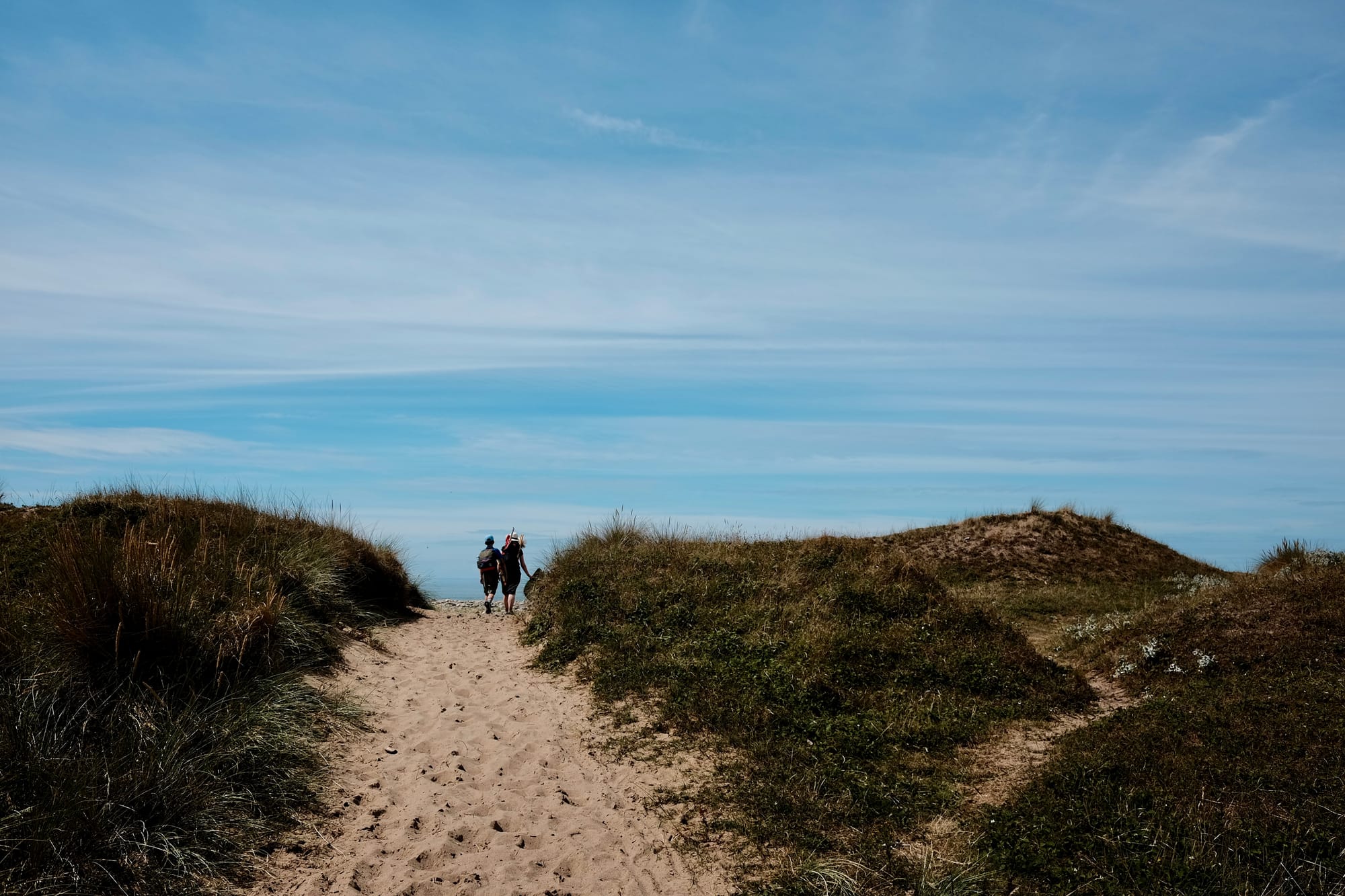 Two people walk on a yellow sandy path through the grass-covered dunes, blue sky above.