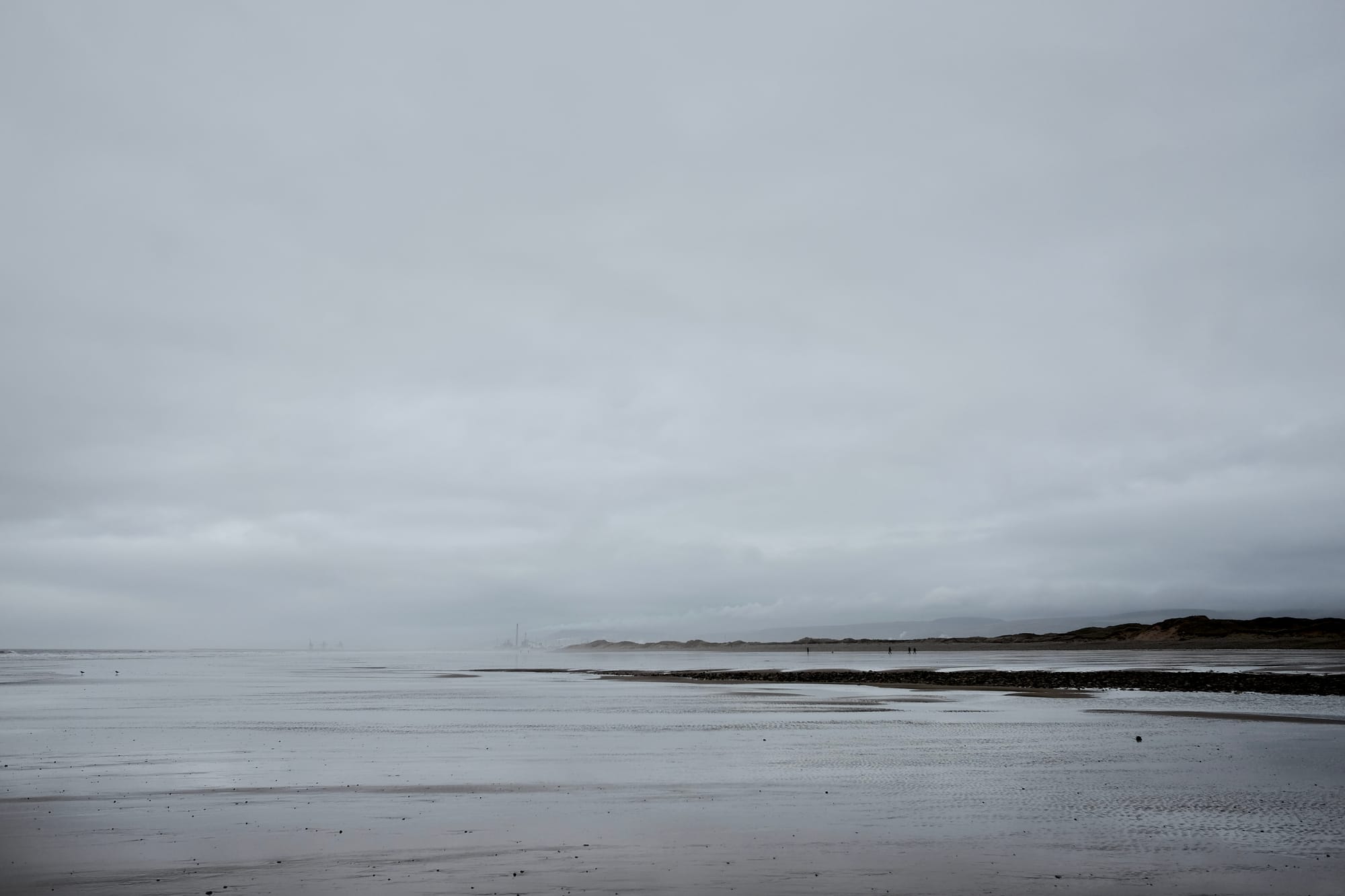 In grey light, distant figures walk on a large swathe of sandy beach covered in water, industrial chimneys in the distance.