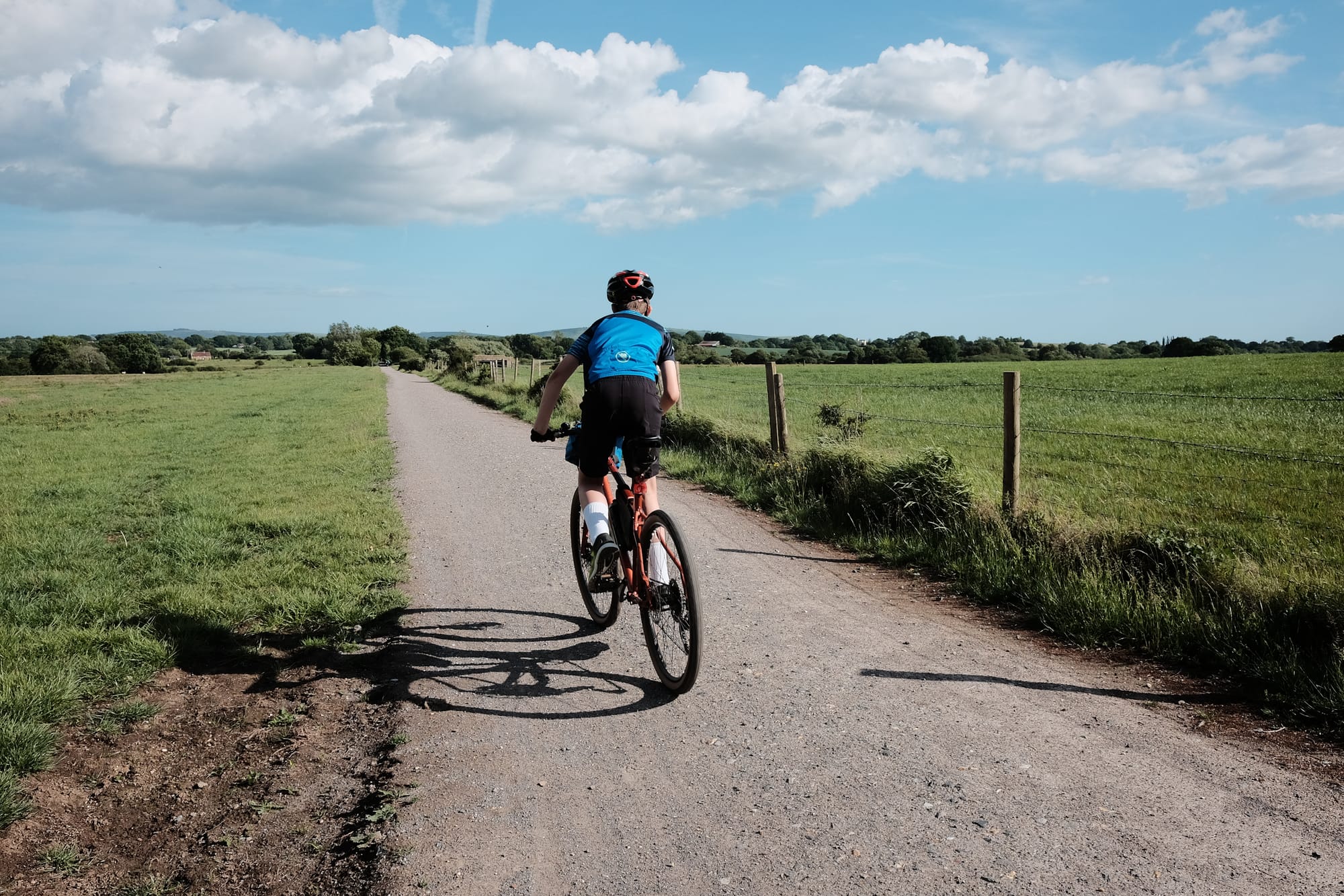 A child in a blue top rides a bike along a grey stone track amidst green fields and beneath a blue sky.