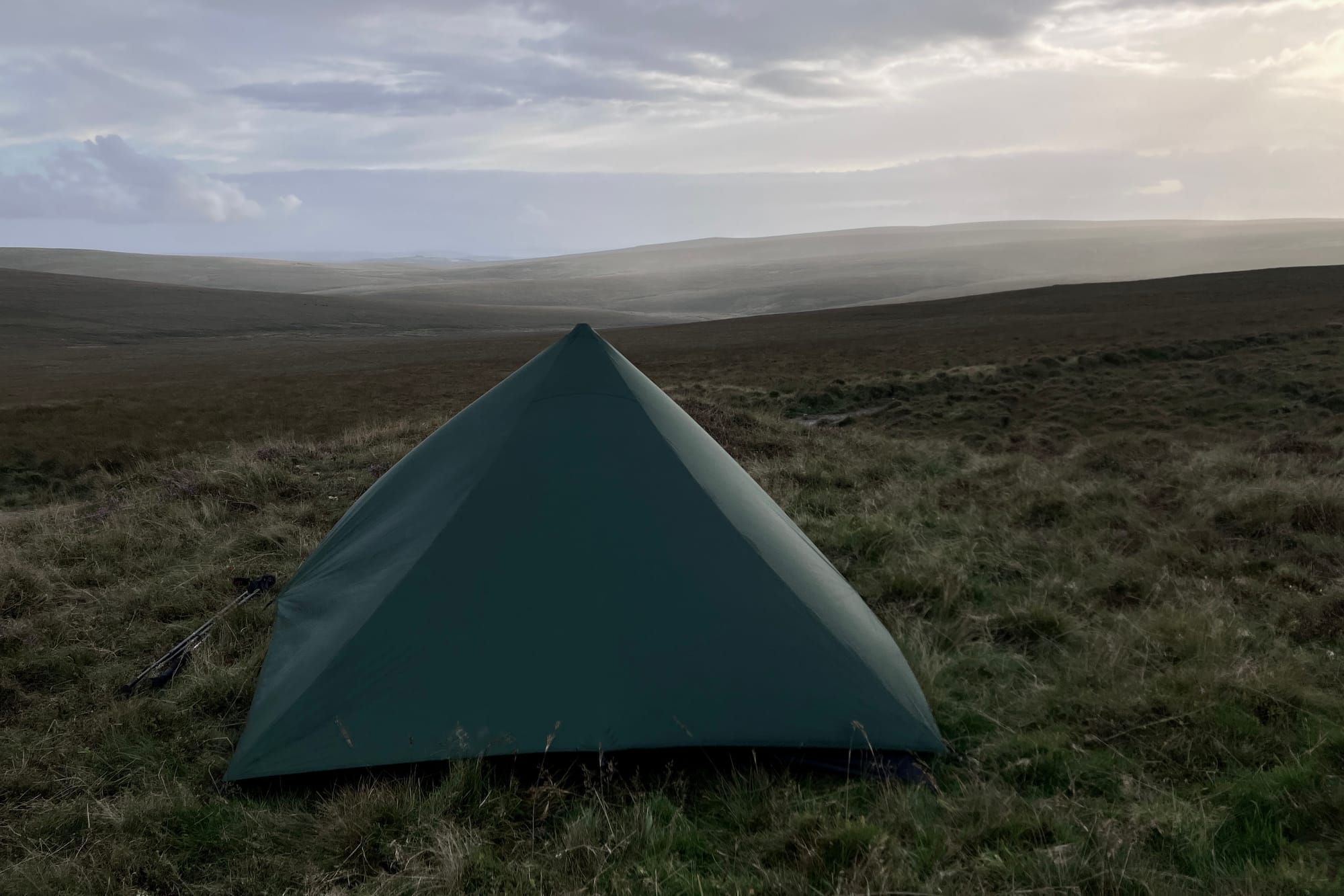 A pyramid shaped green tent pitched in the wide open green space of Dartmoor with the sun breaking through the clouds above