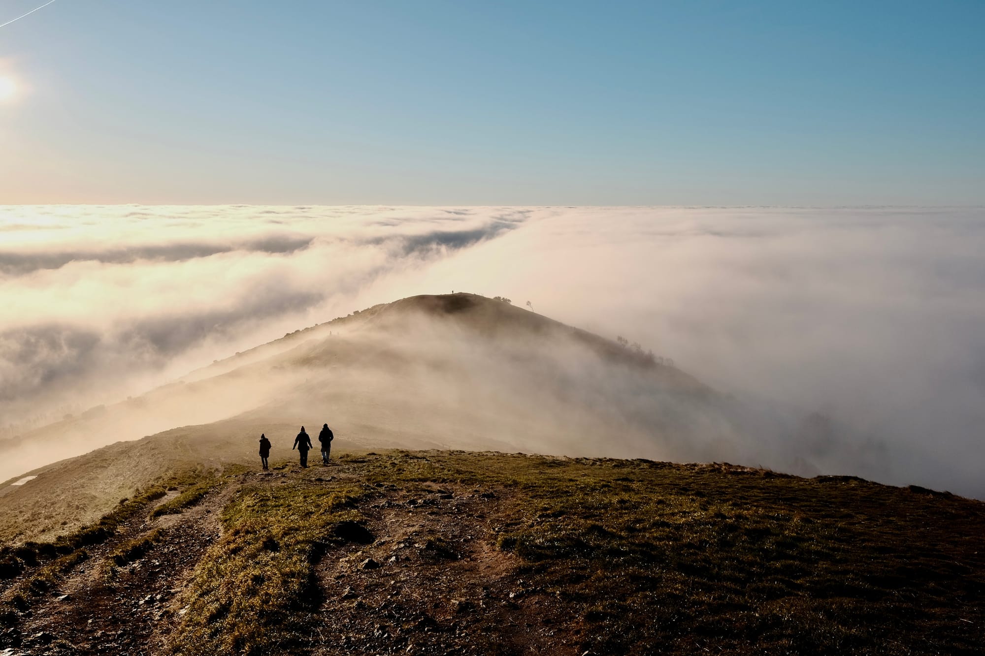 Three walkers proceed along a hilly ridge sticking up into the sunshine above a sea of cloud.