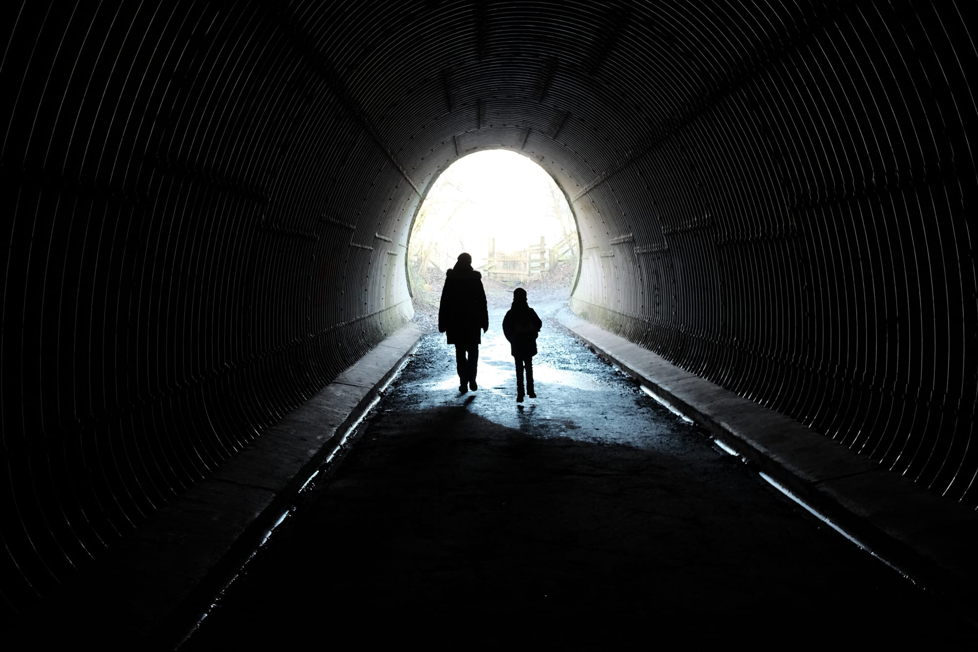 Two figures are silhouetted in a tunnel, a bright light at the far end.