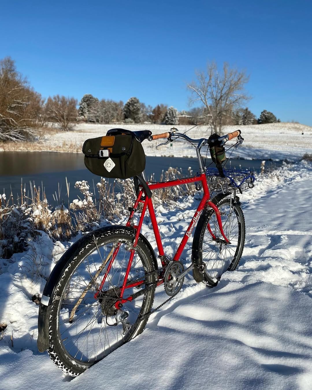 A red bike with a saddle bag and basket stands in deep snow beside a lake.