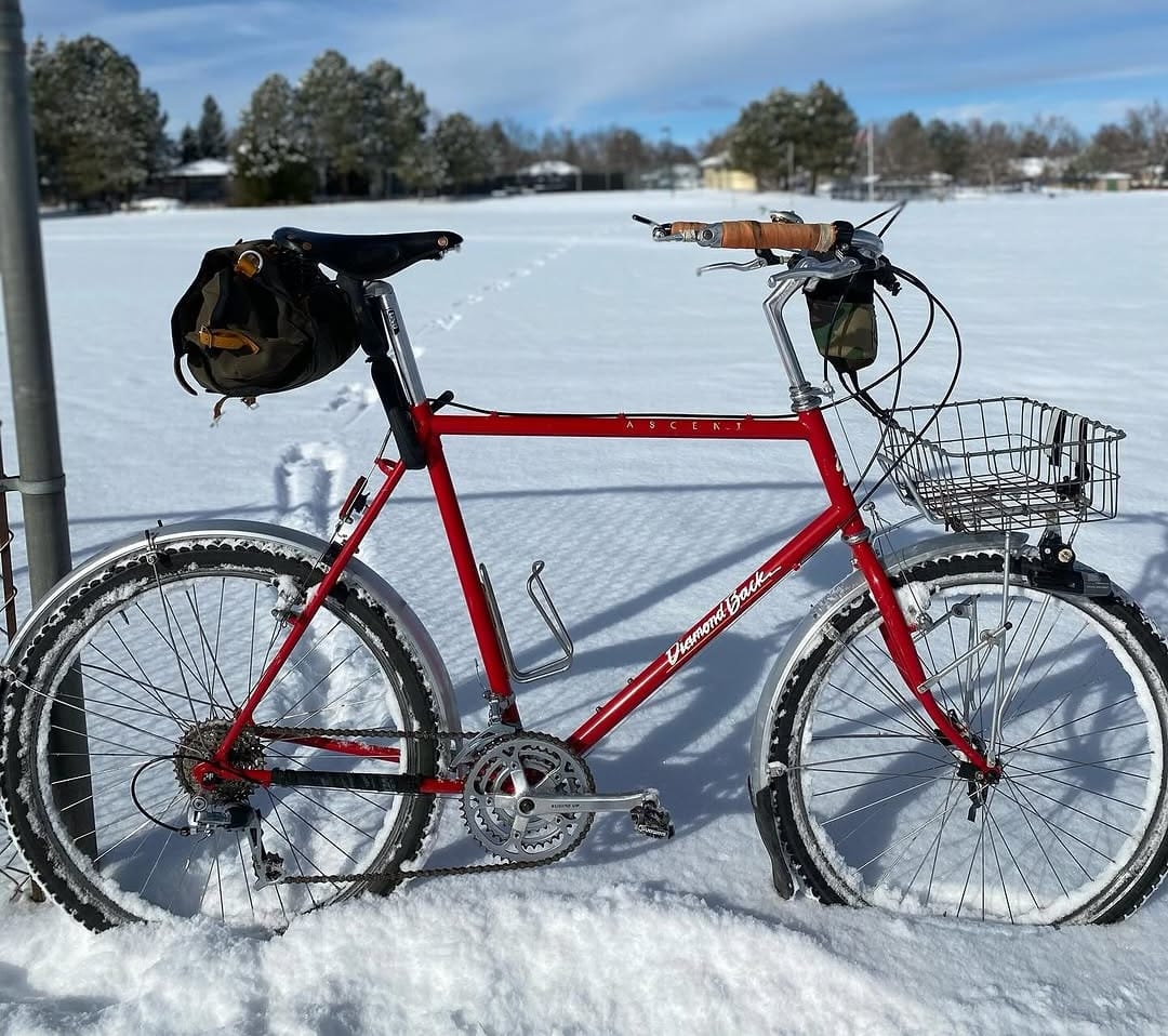 A red bike with saddle bag and basked leans on a fencce with a field of deep snow behind it.
