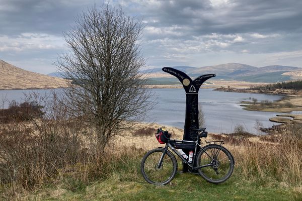 A bike leans on a National Cycle Network waymarker, a tree to one side and a loch in the background.