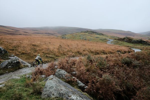 A child cycles along a rough track in high country, grassy slopes on either side.