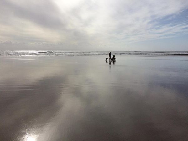 A child on a bike, an adult walking and a dog are silhouetted on the wet sand of Porthcawl at low tide.