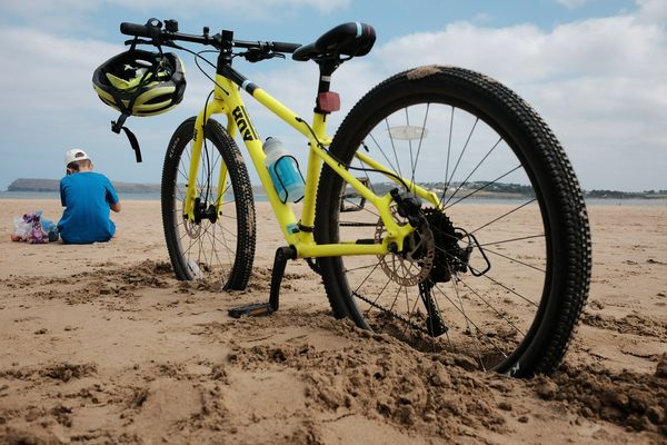 A young boy in a blue t-shirt sits on a sandy beach, his yellow bike propped up in the sand behind him.