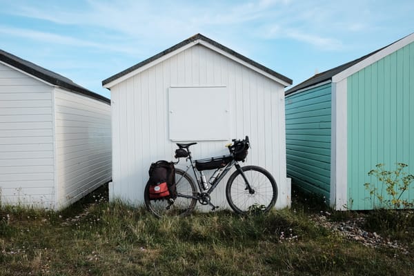 A grey bike with red and black panniers leans on a white beach hut surrounded by grass.