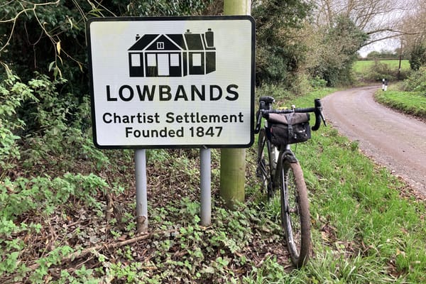 Along a tree-lined coutnry lane, a gravel bike with a black handlebar bag leans on a road sign with the words: "Lowbands: Chartist Settlement Founded 1847"